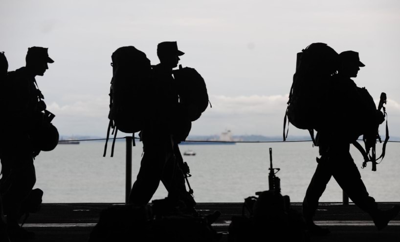 Silhouettes of three service people wearing their kits with water,ships and shoreline in the background. 